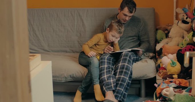 Child learning with dad. They sitting on the sofa at home. Father reading the task and boy writing in the workbook
