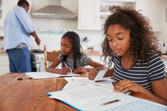 Daughters Sitting At Table Doing Homework As Father Cooks Meal