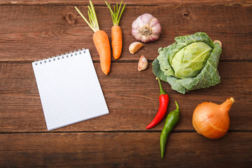 Fresh vegetables with a notepad lie on a wooden background. Cabbage, chili, garlic, carrots and onions on a wooden table. View from above. Space for text.