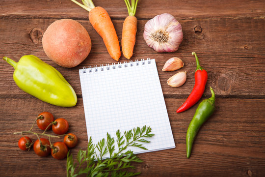 Fresh vegetables with a notepad lie on a wooden background. Bulgarian pepper, chili, potatoes, garlic, carrots and cherry tomato on a wooden table. Space for text.