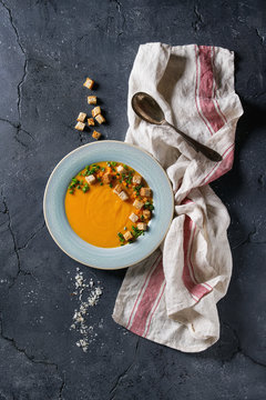 Plate of vegetarian pumpkin carrot soup served with croutons, salt and onion on textile towel over dark texture background. Top view with space