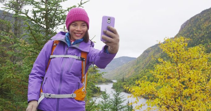 Happy Asian hiker woman taking smartphone selfie at viewpoint in nature fall mountain landscape outdoors. Girl hiking in Autumn forest travel lifestyle. Laurentides, Malbaie, Charlevoix, Quebec Canada