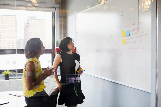 Two women in office, solving problem, using whiteboard, sticky notes stuck on whiteboard