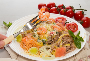 Shrimps on fork above plate. Pasta tagliatelle with grilled shrimps, salmon fish, gouda cheese, serving with tomato cherry, leek and basil. Ingredients around plate with tagliatelle.