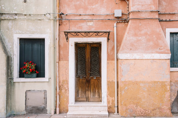 Beautiful colorful house facade on Burano island, north Italy. Partly green partly orange old house wall with faded color. There is old wooden door, pipes and windows with flowers in it
