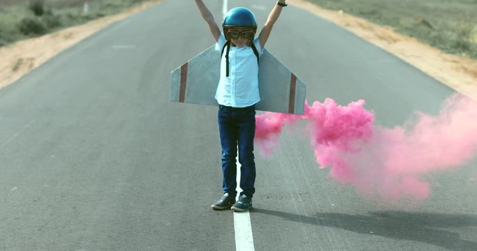 Little boy wearing helmet and styrofoam wings standing on a skateboard on a rural road, pretending to be a pilot. 4K UHD RAW edited footage