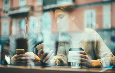Fototapeta na wymiar Young friends using smartphones in coffee bar drinking cappuccino - Co-workers drinking hot coffee in break of work - Window view - Technology and job concept - Focus on woman hand mobile phone