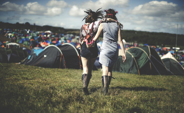 Rear view of two young women at a summer music festival wearing feather headdresses, walking arm in arm towards tents.