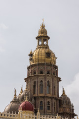 Fototapeta na wymiar Mysore, India - October 27, 2013: closeup of central tower with golden domes of Mysore Palace. Lines of light bulbs , brown stones and more smaller domes against gray sky.