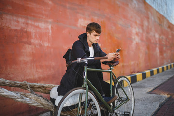 Portrait of boy with brown hair sitting while leaning on bicycle and thoughtfully looking in cellphone. Young man in down jacket and black backpack sitting and using mobile phone with bicycle nearby