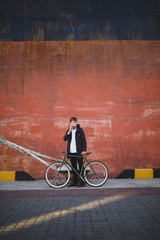Cool boy with brown hair standing with bicycle and looking aside while talking on his cellphone. Young man in down jacket standing and using mobile phone with bicycle nearby