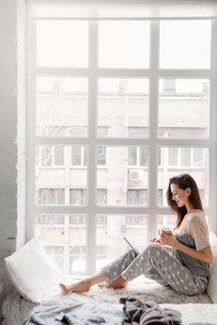 Smiling woman sitting near white window and chatting on laptop. Working start from the morning, making notes, social life, freelance worker concept. Side view picture with free space