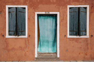 Fototapeta na wymiar Beautiful colorful house facade on Burano island, north Italy. Red old house wall with a door covered with azure cloth and windows