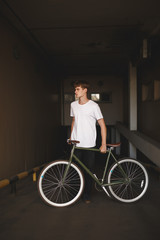 Fototapeta na wymiar Portrait of boy with brown hair standing with bicycle and dreamily looking aside. Photo of young thoughtful man in white t-shirt standing with classic bicycle in parking