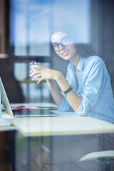 Young woman sitting at office table with laptop,view through window. Young woman