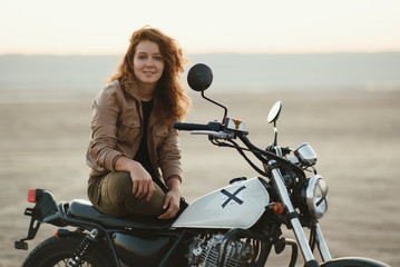 Fototapeta na wymiar young beautiful woman sitting on her old cafe racer motorcycle in desert at sunset or sunrise