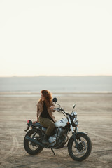 Fototapeta na wymiar young beautiful woman sitting on her old cafe racer motorcycle in desert at sunset or sunrise