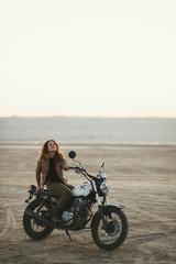  young beautiful woman sitting on her old cafe racer motorcycle in desert at sunset or sunrise