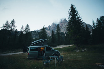 Obraz na płótnie Canvas Small travel vehicle camping van or big car with folding rooftop with bed is parked on secluded wild site under huge mountain formation in dolomites, surrounded by forest