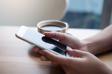 Closeup image of a woman holding and using smart phone with coffee cup on wooden table in cafe