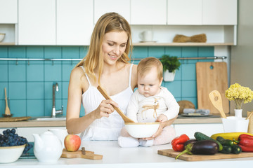 Obraz na płótnie Canvas Young mother is cooking and playing with her baby daughter in a modern kitchen setting. Healthy food concept.