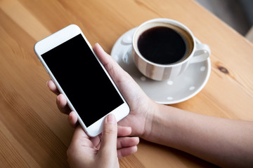 Mockup image of woman's hands holding white mobile phone with blank black screen and white coffee cups in modern loft cafe