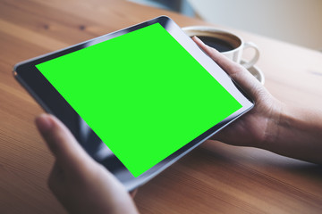 Mockup image of business woman's hands holding black tablet pc with blank green screen and coffee cup on wooden table in cafe background