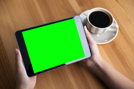 Mockup image of business woman's hands holding black tablet pc with blank green screen and coffee cup on wooden table in cafe background