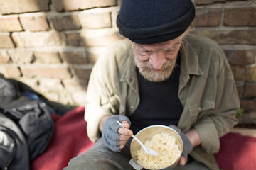 Portrait of homeless old man holding bowl with porridge. Tramp living in the streets of the city,...
