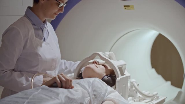 Tracking of cheerful female radiologist putting headphones on woman in disposable gown lying on patient table, then moving her into MRI machine for scan