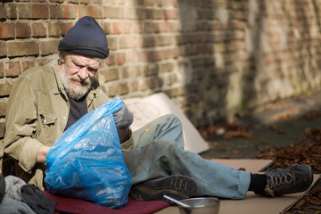 View of a homeless man rummaging in trash bag. Old tramp living in the streets, searching for food...