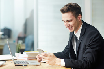 Profile view of smiling young white collar worker texting with his colleague on smartphone while sitting in front of laptop at modern office, waist-up portrait