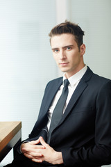 Waist-up portrait of confident young businessman in suit looking at camera while sitting at office desk, white background