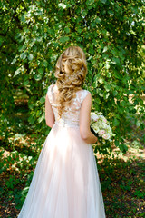 Obraz na płótnie Canvas Beautiful bride in wedding dress with bridal bouquet in the park outdoors, back view. Blond girl with curly hair styling and jewelry. Wedding hairstyle for long hair with stylish hair accessory