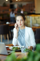 Waist-up portrait of confident young entrepreneur looking at camera with small smile while conducting telephone negotiations at cozy small cafe, blurred background