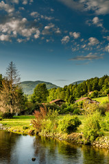 Fototapeta premium river near houses and trees with mountains in back and blue sky