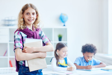 Fototapeta premium Successful schoolboy with copybook and notepad looking at camera in classroom