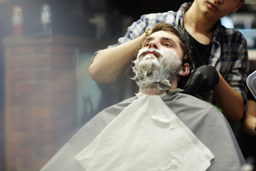 Young client of barbershop with soap foam on his beard and moustache visiting shaving salon