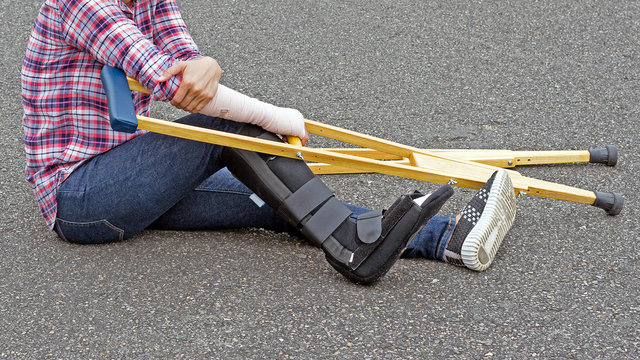 broken leg, injury woman wearing shirt and jeans  with arm splint and black leg splint sitting on floor holding wooden crutches