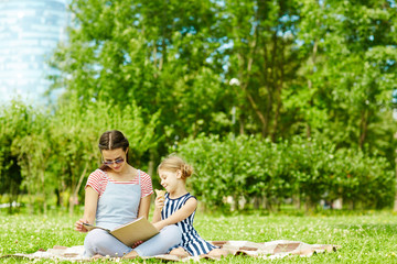 Fototapeta premium Young woman and her daughter reading book of tales on green lawn on summer day