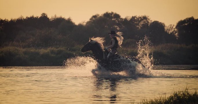 Slow motion 240fps shot of cowboy girl riding horse in water of sunset river
