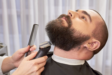 Beard trimming in barbershop. Hands of barber, trimmer and comb.