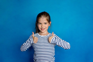 Happy child, little girl showing thumbs up gesture in a striped long sleeves t-shirt  isolated on blue background. Ok sign. Concept of success, school, childhood