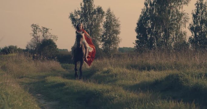 Slow motion 240fps shot of female horserider wearing blue dress and waiving red cloak running at grass field in countryside
