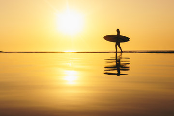 Young woman surfer walking along waters edge with reflection at golden hour