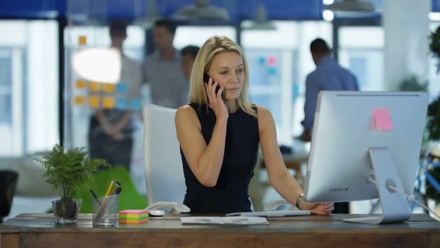 Businesswoman talking on cell phone at her desk in modern office with colleagues in discussion in background.