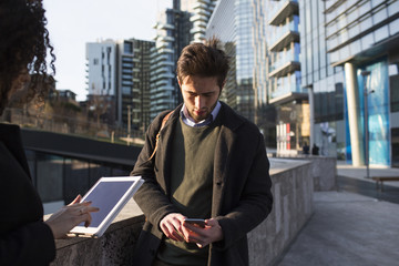 Couple of colleagues using mobile devices outdoors office building