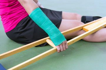 Injured woman wearing sportswear  painful arm with gauze bandage, arm cast and wooden crutches sitting on floor.