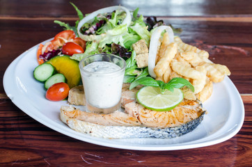 Fish steak, grilled salmon and fresh vegetable salad, french fries and sauce on a white plate