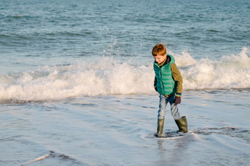 Boy in warm clothes walks along the sea edge with waves behind